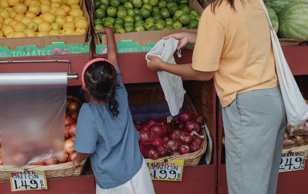 Preschooler reaching for citrus at a produce stall while her mom holds a bag at the Oviedo Farmers Market