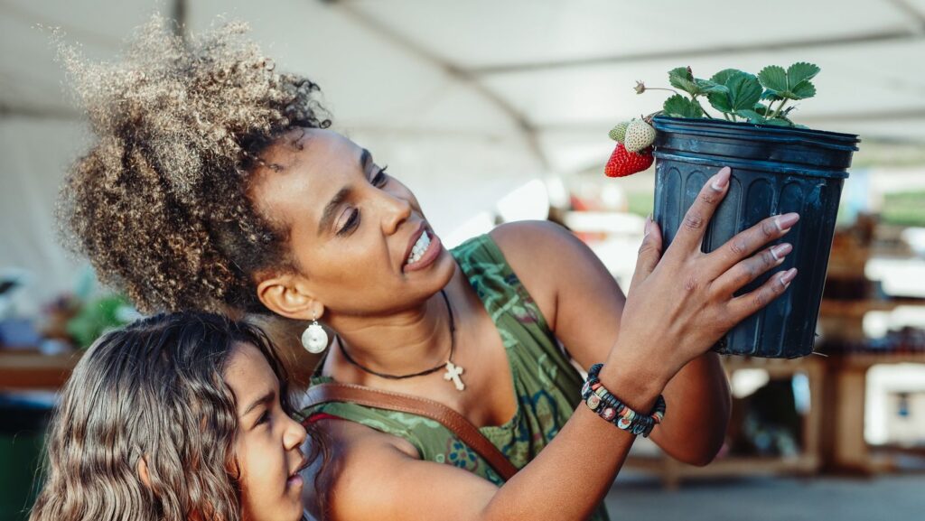 Mom holding up a potted strawberry plant at a farmers market stall while her young daughter looks on