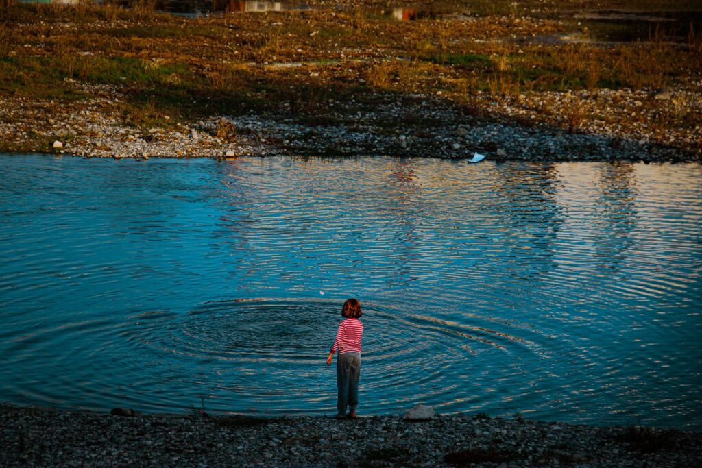 Young child at the edge of a lake watching ripples spread after tossing a stone