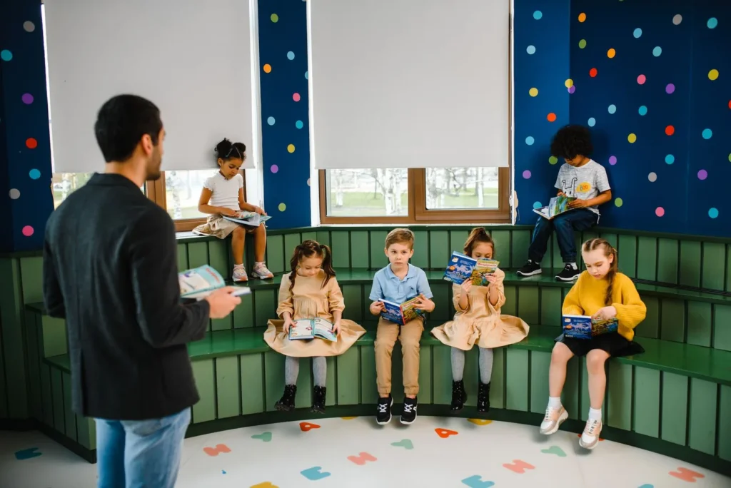 Man reading to six young children seated on a curved green bench in a bright classroom with star-dotted blue wall