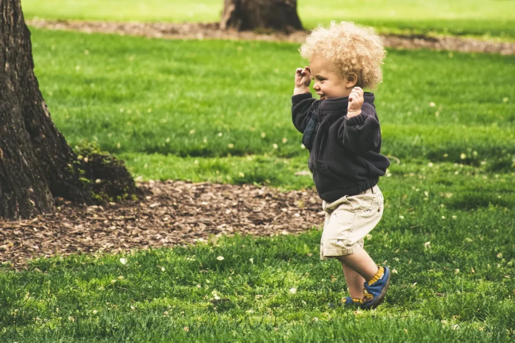 Curly-haired toddler running across green grass near big shady tree trunks in a park