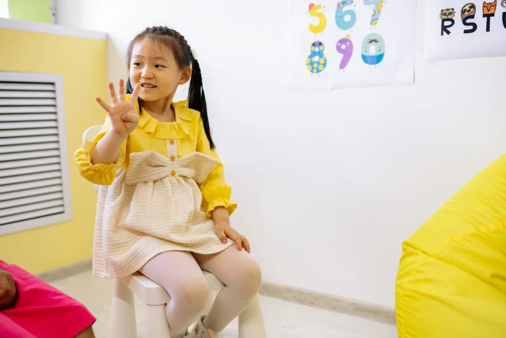 Smiling preschool girl holding up her fingers to count in a bright classroom with number posters behind her