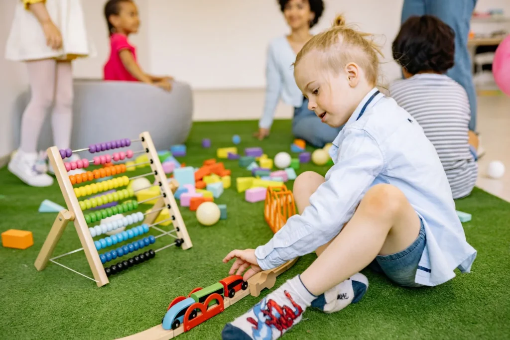 Preschool boy sitting on a green rug rolling toy cars on a track, with an abacus and other children behind him