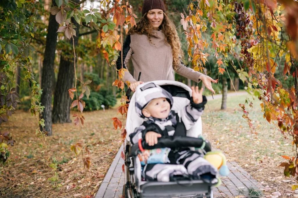 Mother pushing a toddler in a stroller along a tree-lined path with red and gold fall leaves