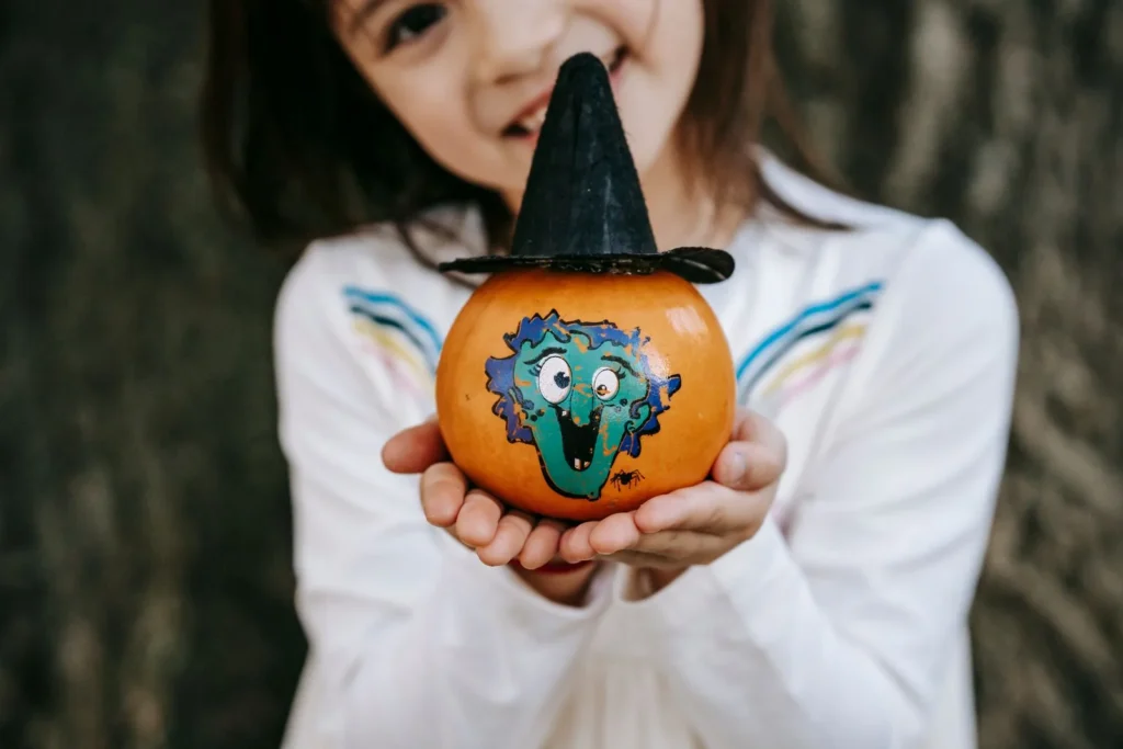 Smiling girl holding out a small hand-painted pumpkin wearing a tiny witch hat