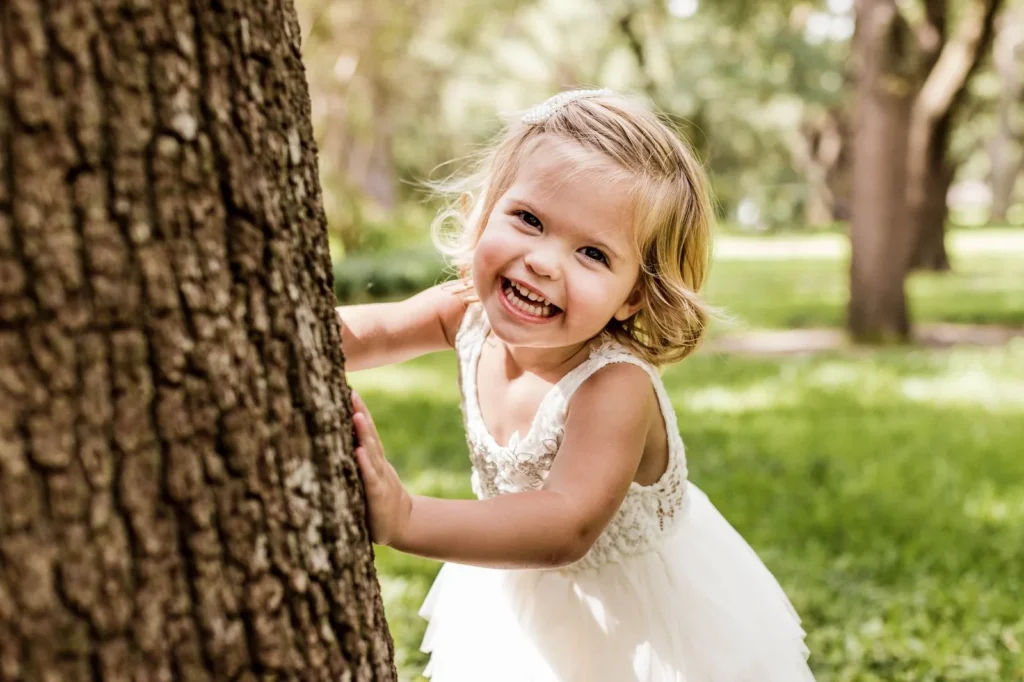 Smiling preschool girl peeking out from behind a big tree trunk in a green park